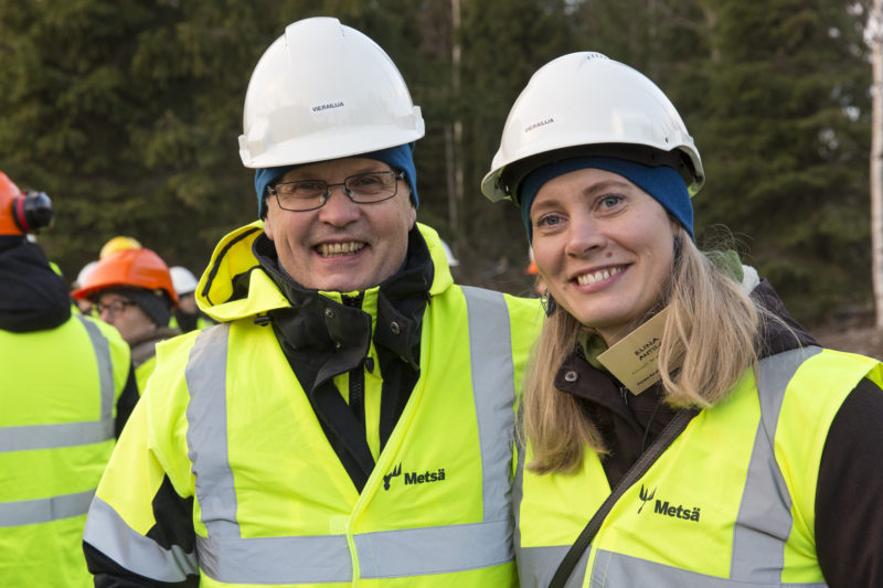 Kaksi iloisen näköistä ihmistä metsässä turvaliivit ja turvakypärät päällään.